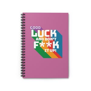May include: A spiral-bound notebook with a magenta cover. The cover features the text "GOOD LUCK AND DON'T F**K IT UP!" in white, with a rainbow-colored design. The notebook has a black spiral binding.