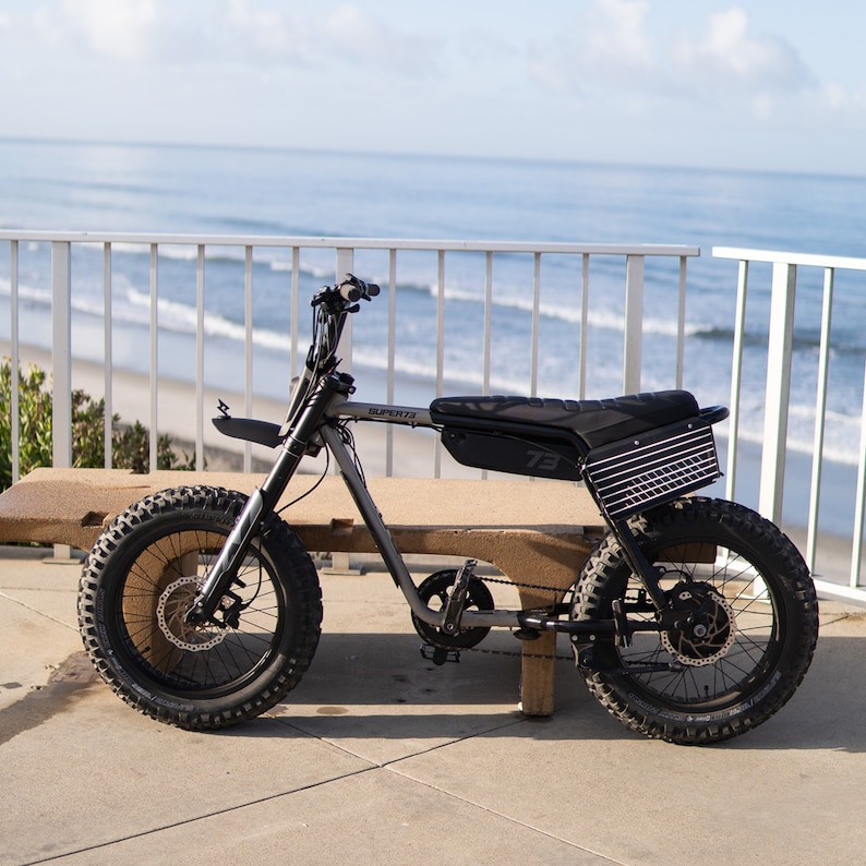 以下が含まれることがあります： 黒とグレーの電気バイクで、大きなノブ付きタイヤを備えています。バイクには黒いシートと、後ろに黒とグレーのバスケットが付いています。バイクは、背景に木製のベンチがあるコンクリートの表面に駐車されています。バイクには側面に「SUPER73」という文字があります。