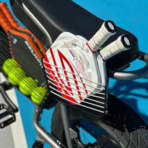以下が含まれることがあります： 銀色の自転車に黒と白のワイヤーラックが取り付けられた白と赤のパドルボールラケット。自転車は黒いシートと黒いタイヤです。