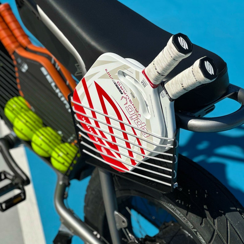 以下が含まれることがあります： 銀色の自転車に黒と白のワイヤーラックが取り付けられた白と赤のパドルボールラケット。自転車は黒いシートと黒いタイヤです。