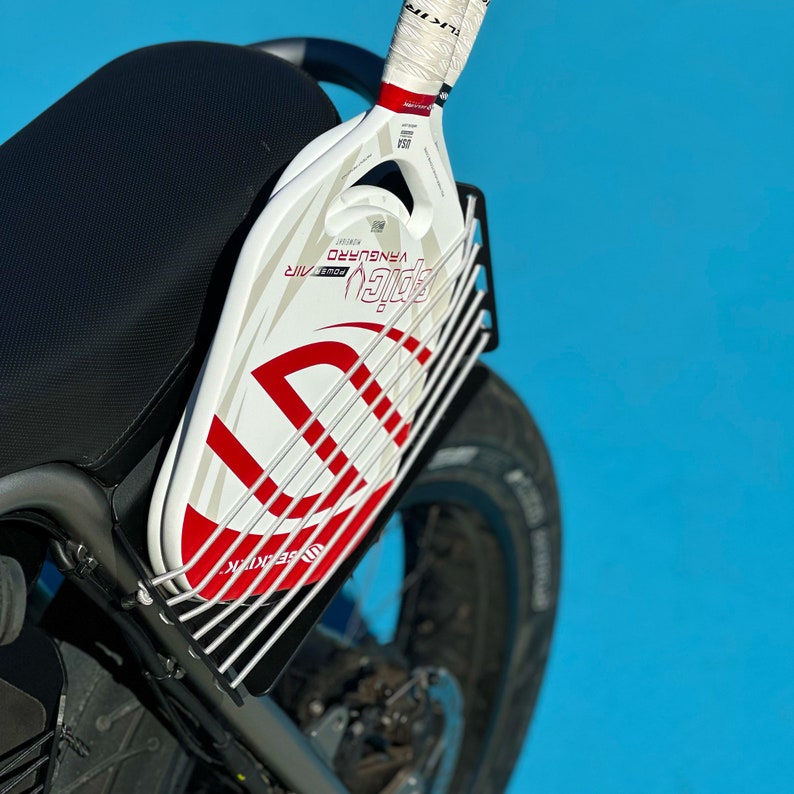 以下が含まれることがあります： 白と赤のピクルボールラケットに「Pickleball Power Air」と「Vanguardo」の文字が書かれています。ラケットは、銀色の金属製のラックが付いたオートバイに取り付けられています。