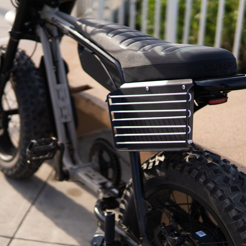 以下が含まれることがあります： 黒い電動自転車。黒いシートと黒い金属製のラックが後ろについています。自転車の後ろには大きな黒いタイヤが付いています。