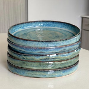 May include: A stack of four round ceramic plates featuring a reactive glaze with shades of blue, teal, and green. The edges of each plate have a dark brown rim, and the surface displays a smooth, glossy finish with subtle color variations.