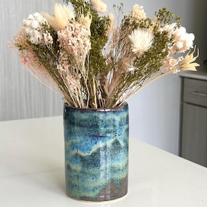 May include: A cylindrical ceramic vase with a glossy blue and green mottled glaze containing a large arrangement of dried flowers in shades of cream, pale pink, and muted green, featuring fluffy bunny tails and various delicate botanical stems.