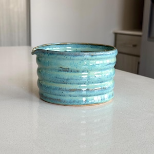 May include: A light blue ceramic pitcher with a textured, wavy design. The pitcher has a rounded shape and a wide opening.