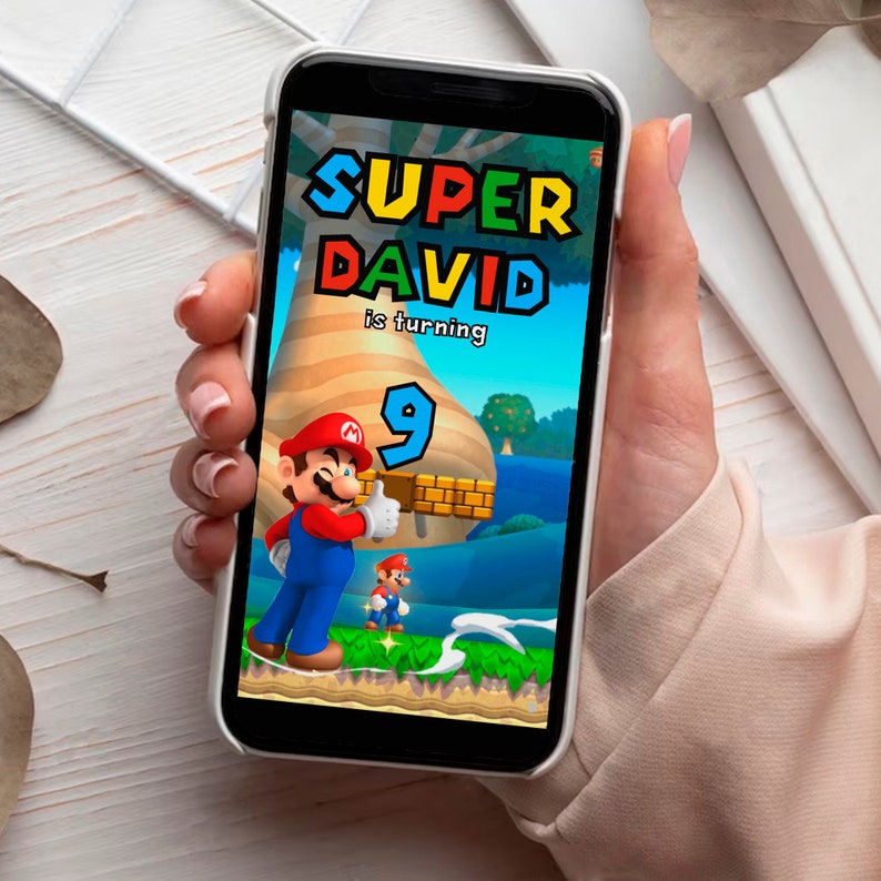May include: A smartphone displays a colorful Super Mario-themed birthday graphic. The text reads "SUPER DAVID is turning 9" with Mario characters and a vibrant landscape. The phone is held in a person's hand.