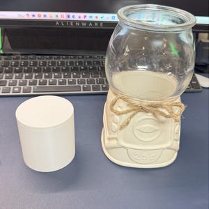 May include: A white ceramic gumball machine with a clear glass globe. The machine is decorated with a jute bow and the text "25¢". A white cylindrical container sits to the left of the machine.