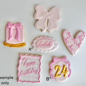 May include: An assortment of decorated sugar cookies in various shapes, including a bow, heart, and rectangle. The cookies are primarily pink and white, with gold accents and text that reads "Leilah" and "happy birthday".