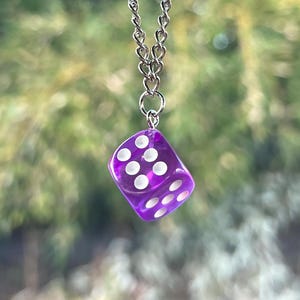 May include: A purple dice car charm hanging from a silver chain. The dice has white dots and is suspended from a car's rearview mirror. The background is a blurred view of green foliage.
