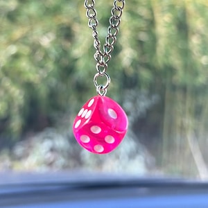 May include: A pink dice car charm hanging from a silver chain. The dice has white dots and is suspended from a car's rearview mirror. The background is blurred, showing green foliage.