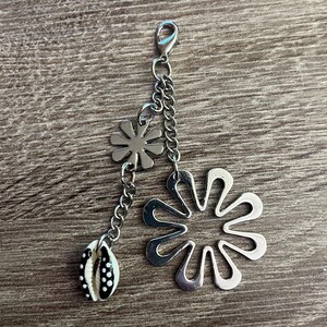 Può includere: Un ciondolo color argento con chiusura a moschettone, catena e tre elementi decorativi. Il ciondolo presenta due pendenti a forma di fiore e una piccola conchiglia con pois bianchi e neri. Il ciondolo è esposto su una superficie di legno.