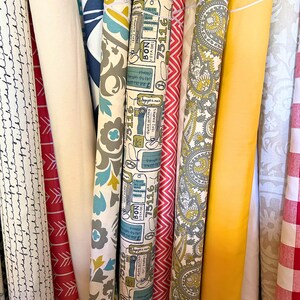 Pu&ograve; includere: Una collezione di rotoli di tessuto con vari motivi e colori. I disegni includono stampe floreali, paisley, chevron e a tema viaggio. I colori vanno dal rosso, bianco e blu al giallo e grigio. Alcuni rotoli hanno il testo "BON VOYAGE 75116".