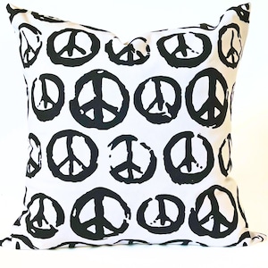 May include: Square white pillow with a repeating pattern of large, black peace symbols. The symbols are irregularly shaped, giving a hand-drawn appearance. The pillow is a decorative item, suitable for a living room or bedroom.
