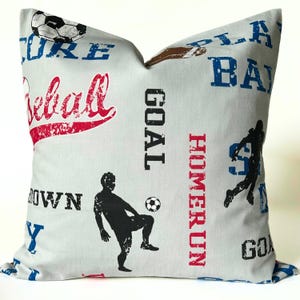 Pu&ograve; includere: Un cuscino decorativo grigio chiaro con testo e grafica a tema sportivo. Il cuscino presenta le parole "Baseball", "Goal" e "Home Run" in rosso e nero. Sono visibili anche sagome di atleti e un pallone da calcio.