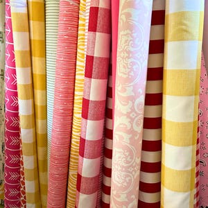 Pu&ograve; includere: Una collezione di rotoli di tessuto con vari motivi e colori. I tessuti includono righe, quadretti e disegni floreali in tonalit&agrave; di giallo, rosso, rosa e bianco. I rotoli sono disposti verticalmente, mostrando la variet&agrave; dei tessuti.