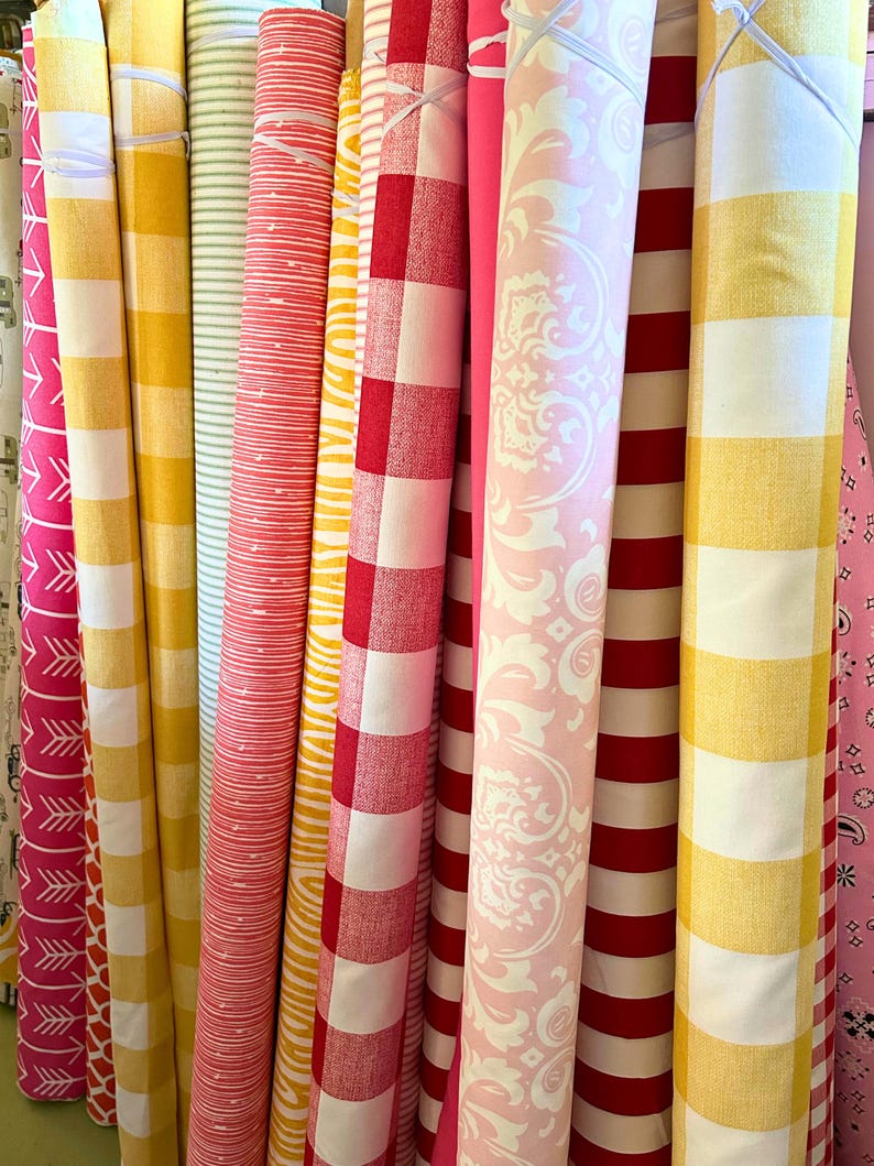 Pu&ograve; includere: Una collezione di rotoli di tessuto con vari motivi e colori. I tessuti includono righe, quadretti e disegni floreali in tonalit&agrave; di giallo, rosso, rosa e bianco. I rotoli sono disposti verticalmente, mostrando la variet&agrave; dei tessuti.