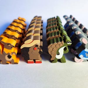 May include: Five colourful animal figurines made of plastic. The figurines are arranged in a row from left to right: a squirrel, a hedgehog, a crocodile, a raccoon, and a mouse. The squirrel is orange, the hedgehog is brown, the crocodile is green, the raccoon is grey, and the mouse is grey with blue accents.