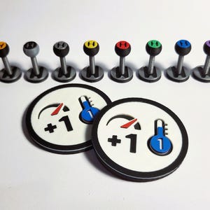 以下が含まれることがあります： 黒い縁取りのある白い丸いゲームピースが2つ。それぞれのピースには、+1 を指す赤い針と青い数字 1 が付いた青い温度計があります。これらのピースはボードゲームで使用されます。