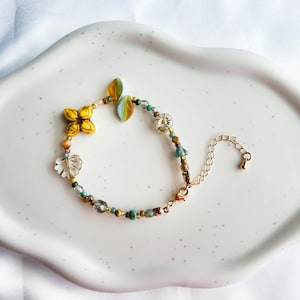 Puede incluir: Un delicado brazalete dorado con una cadena de cuentas que presenta una variedad de cuentas de vidrio de colores, incluyendo amarillo, azul, verde y blanco. El brazalete tiene un dije floral y un dije de hoja.
