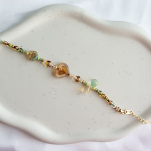 Puede incluir: Un delicado brazalete de cadena dorada con cuentas de vidrio verde y marrón y un dije en forma de corazón. El brazalete está sobre un plato de cerámica blanco.