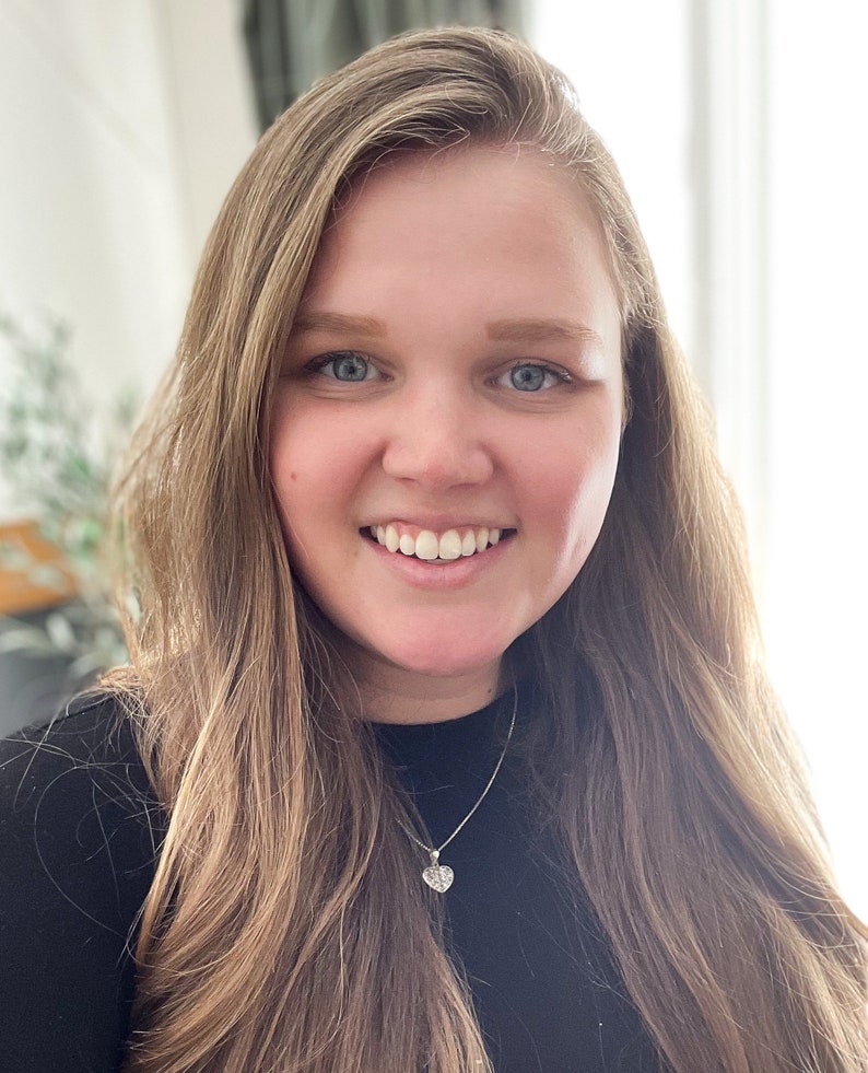 May include: A woman with long, light brown hair is smiling at the camera. She is wearing a black shirt and a silver necklace with a heart pendant.