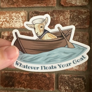 Puede incluir: Una cabra blanca con un gorro de marinero está en un pequeño bote de madera sobre agua azul. El bote tiene un remo de madera. El texto "Whatever Floats Your Goat" está debajo del bote.