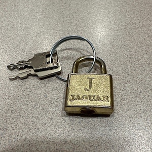 May include: A gold padlock with the letter "J" and the word "JAGUAR" engraved on the front. The padlock is attached to a silver key ring with two keys.