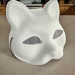 May include: A white cat mask with mesh eye coverings and a black string. The mask is made of a smooth, solid material and is designed to fit the face. The cat mask is a costume accessory.