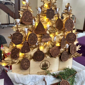May include: Wooden Christmas ornaments with various religious and nature-themed designs, displayed on a decorative wooden tree. The ornaments are brown with leather cords. Some ornaments have text like "Peace on Earth" and "Silent Night".