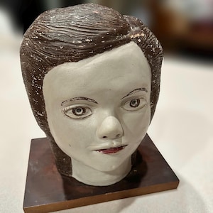 May include: A ceramic bust of a person with brown hair and a white face. The bust is painted with brown and red details. The bust is sitting on a wooden base.