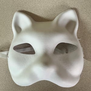 May include: A white cat-shaped mask with eye cutouts and attached elastic straps. The mask has a smooth surface and is designed to be worn on the face. The ears are pointed and the nose and mouth are subtly defined.