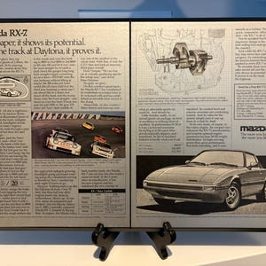 May include: Framed vintage Mazda RX-7 advertisement. The ad features text, a diagram of the car's engine, and a photo of the car on a racetrack. The Mazda logo is visible.