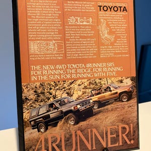 May include: A framed advert for the Toyota 4Runner SR5, showing two vehicles on a dirt track. The ad copy highlights the vehicle's features, including its 4-wheel drive and spacious interior. The text "4RUNNER!" is prominently displayed.