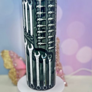 May include: A stainless steel tumbler with a black and silver design featuring a variety of wrenches and screwdrivers. The tumbler has a clear lid and a stainless steel straw.
