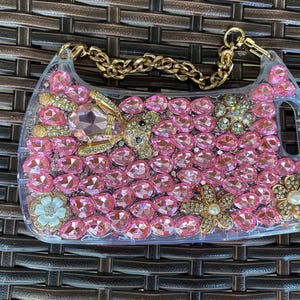 May include: Clear, rectangular phone case adorned with pink rhinestones and gold accents. The case features a gold chain strap and decorative elements including a teddy bear, flowers, and a pearl.