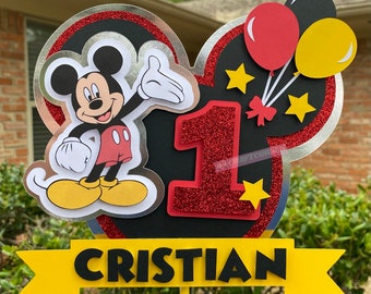 May include: A red, black, and yellow Mickey Mouse themed cake topper with a red glitter number one, a Mickey Mouse character, and the name "Cristian" on a yellow banner.