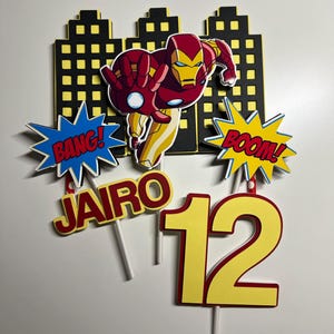 Könnte beinhalten: Iron Man Kuchentopper-Set mit einer roten und gelben Iron Man Figur, einer schwarzen Skyline, einer roten und gelben Sprechblase "BANG!", einer roten und gelben Sprechblase "BOOM!", einem roten und gelben Namenstopper "JAIRO" und einem gelben Zahlenkuchentopper "12".