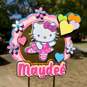 Puede incluir: Adorno para tarta en capas con Hello Kitty con un atuendo de bailarina rosa, rodeada de flores y corazones rosas y azules. El nombre "Maydee" se muestra en letras rosas y blancas. El adorno se sujeta con dos palitos dorados.
