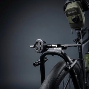 Op de afbeelding: Een zwarte bagagedrager voor een fiets met een ronde montagebeugel met een centrale kruisvormige gleuf. De drager is voorzien van de witte tekst tailfin en is bevestigd aan een fietsframe met een olijfgroene zadeltas met het label CXWXC.