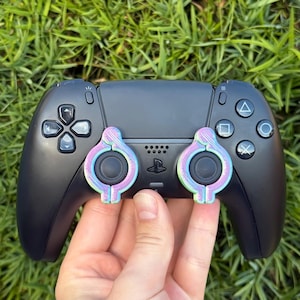 Puede incluir: Un mando de PlayStation 5 negro con dos agarres para el pulgar iridiscentes de color rosa y verde.