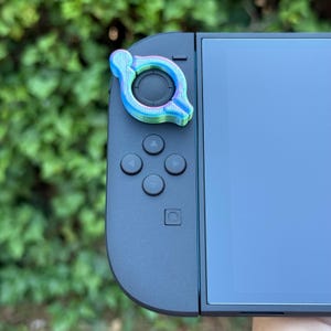 以下が含まれることがあります： 黒い携帯型ゲーム機に取り付けられた、マルチカラーのコントローラーアクセサリー。 アクセサリーは、青、緑、ピンクの色合いを持つ円形のデザインです。 デバイスには画面といくつかのボタンがあります。