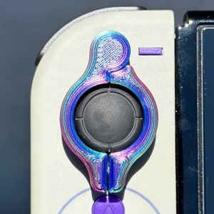 May include: A close-up of a iridescent purple and blue gaming button with a black center. The button is attached to a white controller.