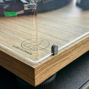 Puede incluir: Primer plano de un tocadiscos de madera con una cubierta de acrílico transparente. El tocadiscos tiene un acabado de veta de madera marrón claro y un logotipo plateado de Audio-Technica. Un cierre de metal negro asegura la cubierta.