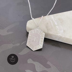 Puede incluir: Un collar de tono plateado con un colgante hexagonal que presenta un borde grabado estilo rayos de sol y la letra R en una fuente de estilo gótico en el centro. El colgante cuelga de una delicada cadena de eslabones de tono plateado.