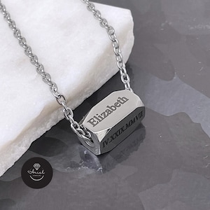 Op de afbeelding: Een zilverkleurige ketting met een geometrische hanger. De hanger is gegraveerd met de naam "Elizabeth" en Romeinse cijfers. De ketting wordt getoond op een wit oppervlak met een donkergrijze achtergrond.