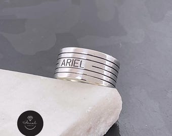 Puede incluir: Un anillo de plata con el nombre "ARIEL" grabado. El anillo tiene un diseño texturizado y rayado.