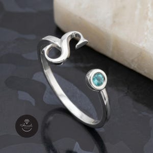 Könnte beinhalten: Ein silberner Ring mit offenem Design, der den Buchstaben "S" und einen runden, hellblauen Edelstein zeigt. Der Ring ist vor einem dunklen, strukturierten Hintergrund mit einem weißen Stein im Hintergrund platziert.
