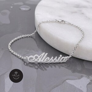 Könnte beinhalten: Ein Namensarmband aus silberfarbenem Metall mit dem Namen Alessia in Schreibschrift. Das Armband besteht aus einer feinen Gliederkette mit Karabinerverschluss und weist eine polierte Oberfläche auf, die das Licht reflektiert.