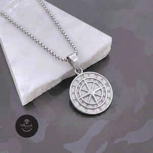 Può includere: Una collana con pendente color argento con un design a bussola. Il pendente rotondo ha una stella al centro ed è circondato da testo. La catena della collana è a maglia scatolata. Il pendente è esposto su una superficie bianca e angolata.