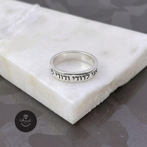 May include: A silver band ring featuring black engraved Hebrew text that reads "אני לדודי ודודי לי" around the outer surface. The ring has a smooth, polished finish and a consistent width, showcasing clear, dark lettering against the metallic silver surface.